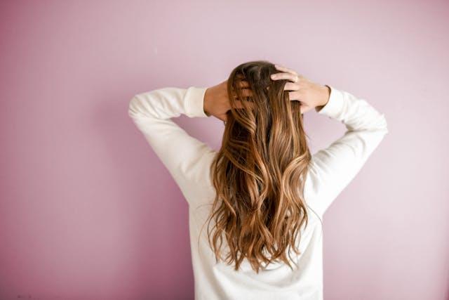 femme de dos avec pull blanc et des cheveux longs brillants qui se touche les cheveux sur fond rose