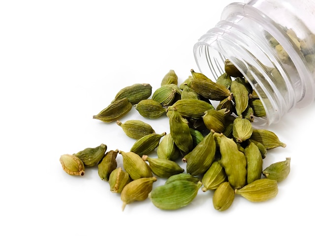transparante flessenhals op een witte achtergrond met groene kardemomzaadjes die uit de pot komen