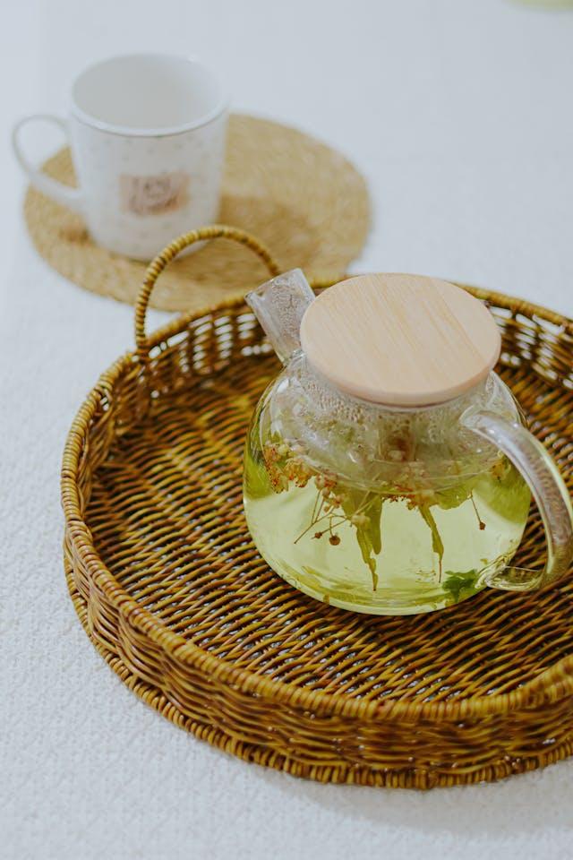 Théière avec des fleurs de tilleul dans un plateau rond en rotin avec en arrière-plan une tasse blanche posée sur un set rond beige
