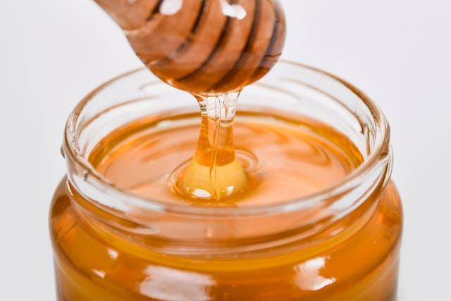 Cuillère en bois ronde enduite de miel qui coule dans un pot transparent rempli de miel