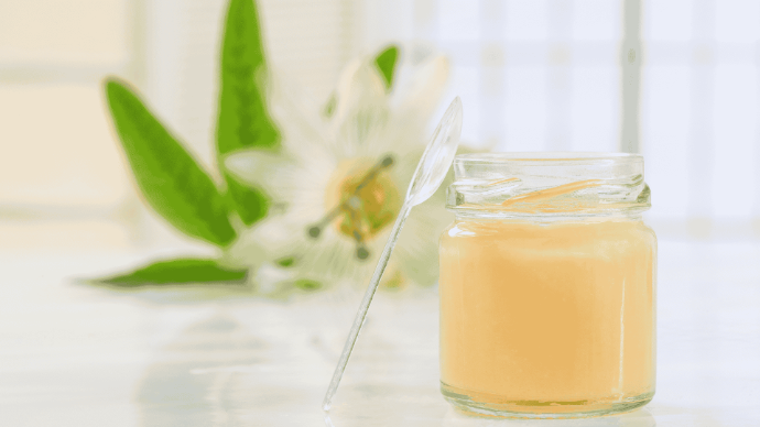 Pot de gelée royale avec cuillère transparente en plastique debout à côté et fleur blanche avec feuilles vertes en arrière-plan