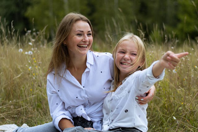 Maman en chemise blanche assise dans des herbes hautes qui sourit avec sa fille qui tend un bras à côté d'elle