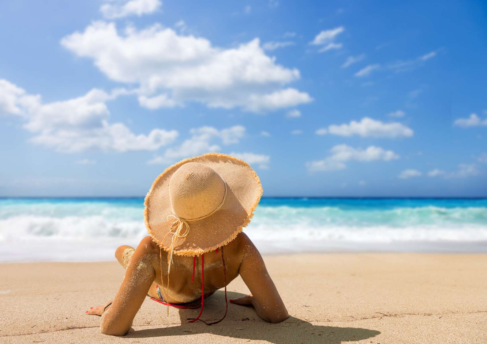 Femme de dos en maillot de bain allongée sur les coudes avec un chapeau de paille face à la mer