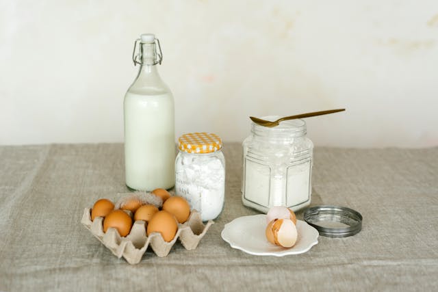 Bicarbonate de soude dans pots de confiture avec boîte d'œufs, œuf cassé dans assiette blanche et bouteille en verre de lait posés sur nappe en lin 