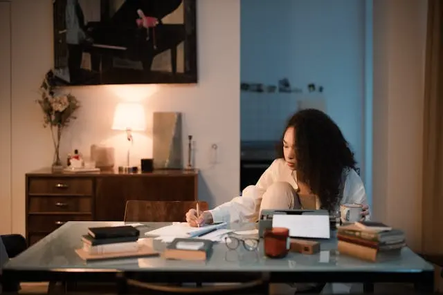 Jeune femme noire concentrée qui écrit sur un bureau sur une table avec livre et machine à écrire dans une salle à manger