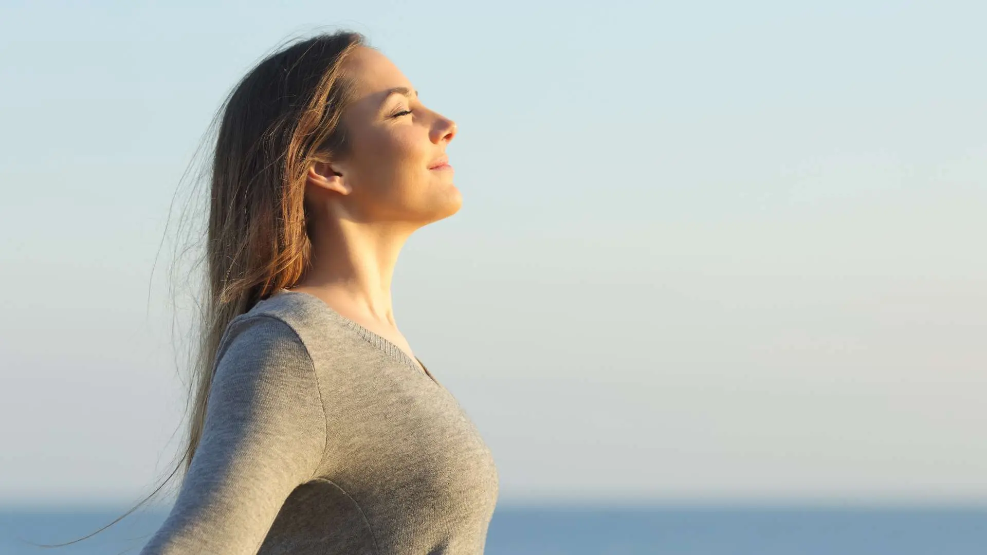 Jeune femme qui respire les yeux fermés la tête levée en souriant avec mer et ciel bleus en arrrère-plan
