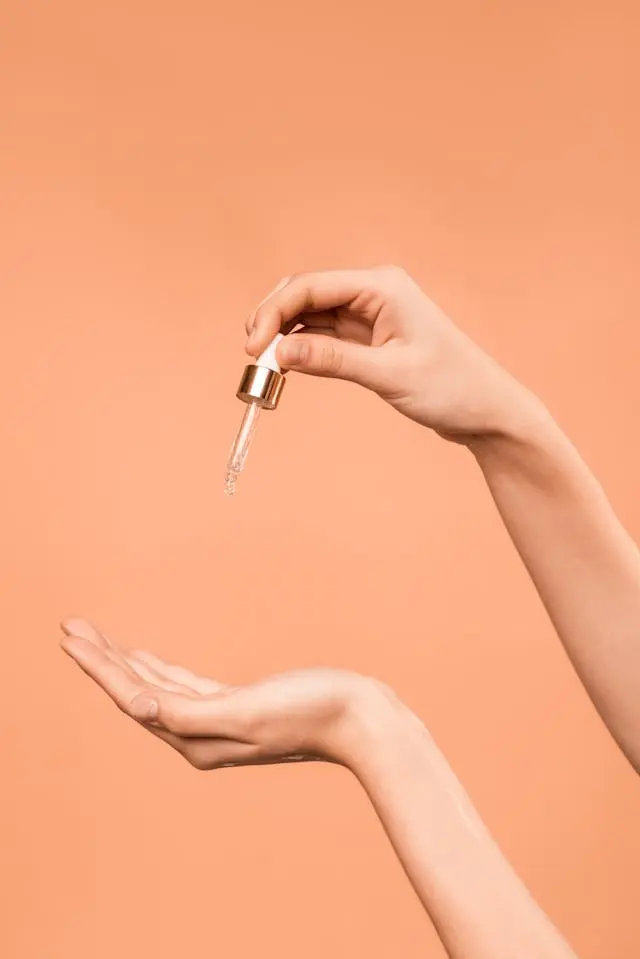 Avant-bras de femme de profil avec une main qui tient une pipette d'huile végétale au-dessus de l'autre sur fond corail